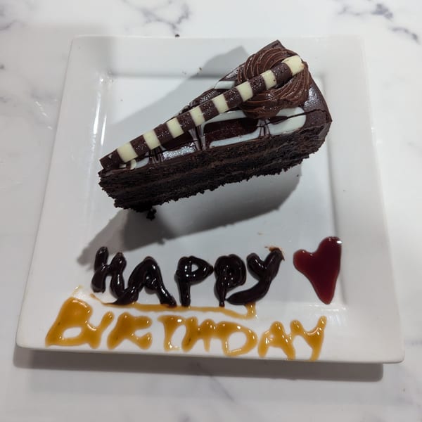 A slice of chocolate cake with "Happy Birthday" written in icing on a white plate.