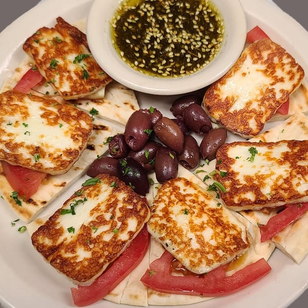 Grilled halloumi cheese, pita bread, sliced tomatoes, olives, and a dipping sauce with sesame seeds.